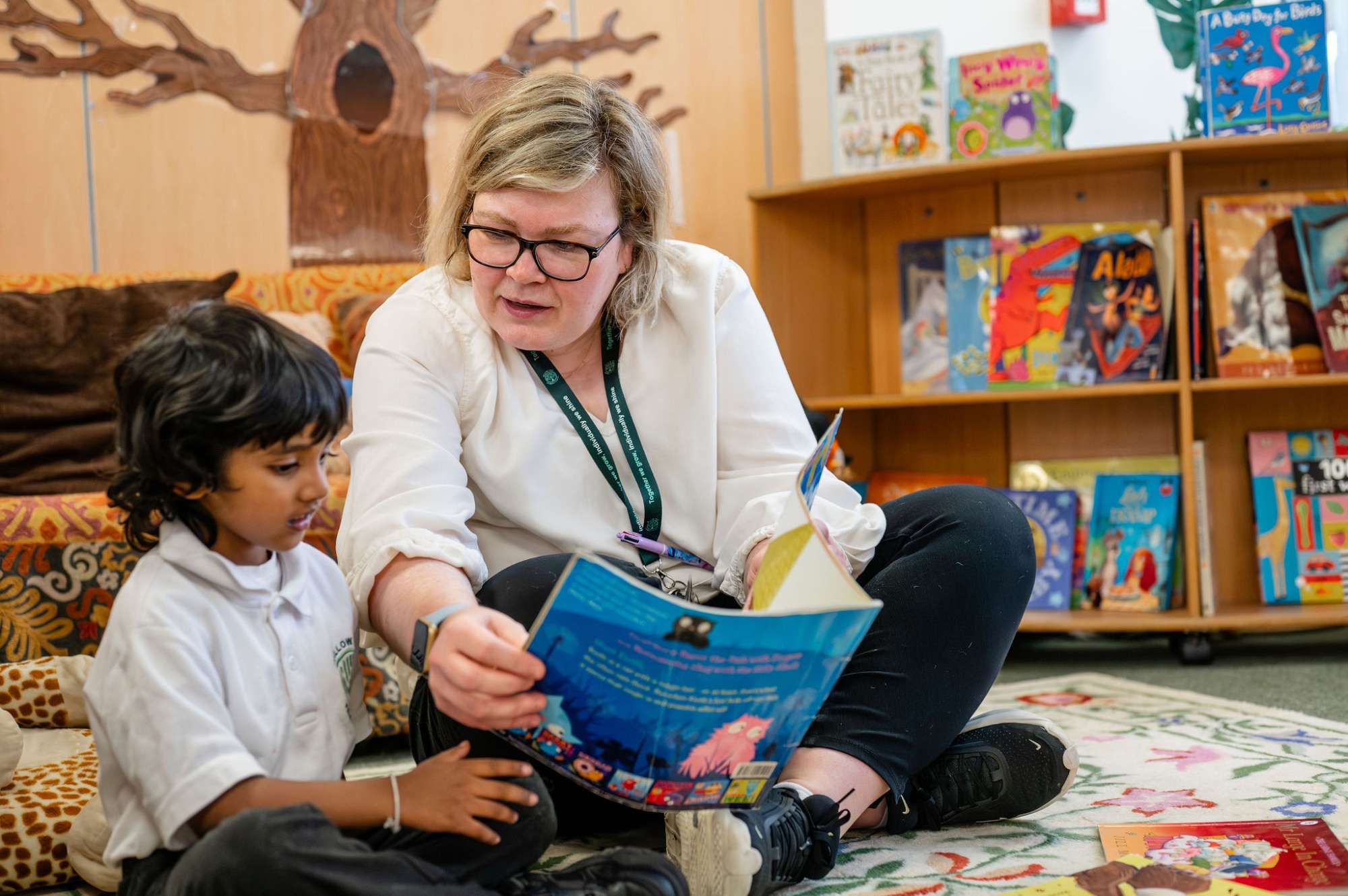 A member of staff reads with a pupil cross-legged on the floor in the library
